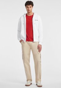 Bijela zip-up majica preko crvene majice, uparen s krem hlačama s bočnim džepovima. Model nosi bijele i bež tenisice.