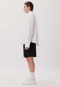 Camicia bianca oversize con maniche lunghe, abbinata a pantaloni corti blu navy e sneakers bianche. Tessuto morbido, design semplice e colletto con bottoni.