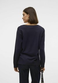 Pull bleu marine à encolure dégagée, avec des poignets et un ourlet côtelés. Texture lisse, silhouette ajustée, associé à un jean foncé et un sac à main.