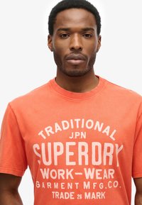 Mand iført en orange t-shirt med hvid tekst, der lyder "Traditional Superdry Work-Wear Garment MFG. Co. Trade Mark" på hvid baggrund.