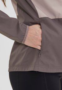Vandtæt jakke i lysegrå og taupe, med en lynlåslomme i siden, lange ærmer og en let struktureret overflade.