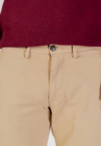 Pantalon en coton beige à finition texturée, doté d'une fermeture à boutons sur le devant et de passants pour ceinture, associé à un pull en maille bordeaux.