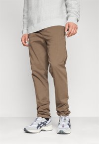 Jasnoszary sweter o teksturze, brązowe spodnie slim-fit oraz białe sneakersy z szarymi i czarnymi akcentami. Skupienie na materiałach i prostym designie.