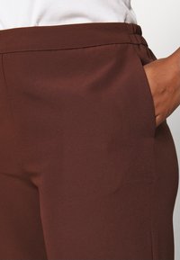 Brązowe spodnie z gładkiej tkaniny o subtelnym połysku, z elastycznym pasem i bocznymi kieszeniami. Krój jest dopasowany, zapewniający strukturalne wykończenie.