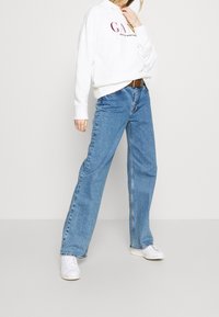 Sudadera blanca con logotipo, vaqueros de pierna ancha en azul claro y zapatillas blancas. Material de algodón, corte casual, con un detalle mínimo.