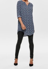 Tunic blu navy con motivo circolare, abbottonata al collo, abbinata a leggins in pelle nera e scarpe con tacco alto nere.