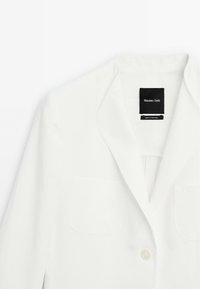 Blazer bianco su misura con rever a taglio, due tasche frontali, chiusura con un solo bottone e etichetta nera del marchio all'interno del colletto.