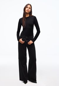 Fekete hosszú ujjú body, testhez simuló szabással, magas derekú, bő szárú fekete farmernadrággal párosítva. A modell zsebébe tett kézzel áll.