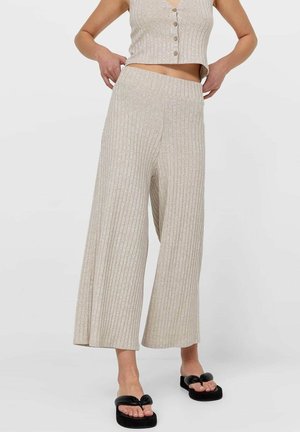 Kvinde iført beige vidde ribbestrikkede bukser, matchende ærmeløs knapdet crop top og sorte åbne flade sandaler med sløjfedetalje.