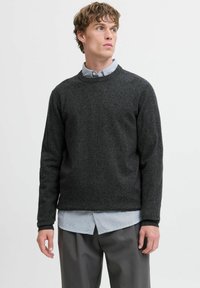 Homme portant un pull gris foncé par-dessus une chemise à rayures claires et un pantalon gris, regardant sur le côté devant un fond uni.