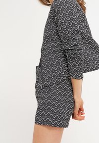 Frau, die ein Blazer mit schwarz-weißem Zickzackmuster und einen passenden Rock trägt, steht mit entspannten Armen an ihrer Seite vor einem schlichten Hintergrund.