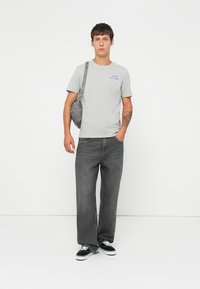 Giovane uomo che indossa una maglietta grigia con grafica, jeans grigio scuro, sneakers nere, con una borsa a tracolla grigia e un tatuaggio sul braccio sinistro, che sta in piedi contro un muro bianco.