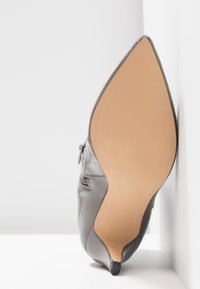 Chaussure à talon haut en cuir gris à bout pointu, debout, montrant la semelle beige lisse et le petit embout de talon noir.
