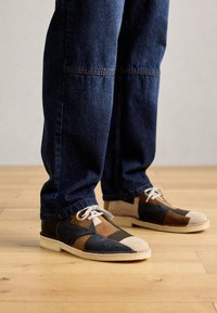Person wearing dark blue jeans and multi-colored patchwork leather shoes with white soles standing on wooden floor.
