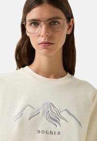 Cremefarvet sweatshirt med sølvfarvet bjergbroderi og "BOGNER"-tekst. Modellen bærer oversized briller med metalramme.