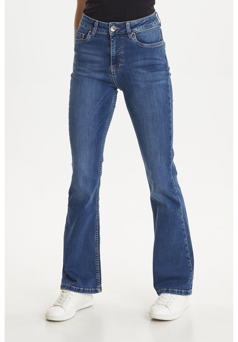 Personne portant un jean bootcut bleu taille mi-haute et des baskets blanches, debout devant un fond clair uni.