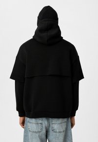 Sweatshirt noir avec capuche, présentant un design superposé et des manches larges. Associé à un jean en denim clair. Tissu simple et texturé.