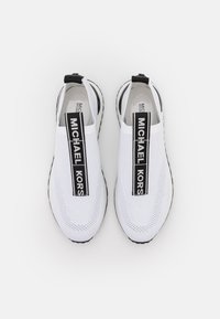 White slip-on sneakers with a knitted mesh upper, black branding strap, and textured rubber sole. Minimalist design with no visible laces.