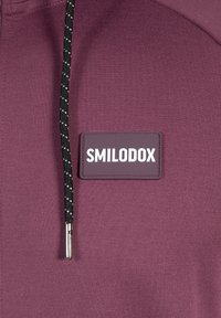 Bordeauxfarvet hættetrøje med en glat tekstur, justerbar sort snor med hvide detaljer og et fremtrædende Smilodox logo.