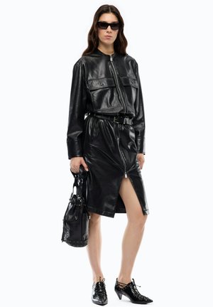 Donna che indossa giacca di pelle nera e gonna con spacco frontale con zip, che tiene una borsa nera, scarpe nere con tacco e occhiali da sole scuri.