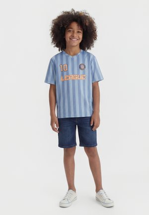 Barn med krøllet hår iført en blå stribet sportstrøje med "10" og "LEAGUE" tekst, denimshorts og hvide sneakers, smilende.