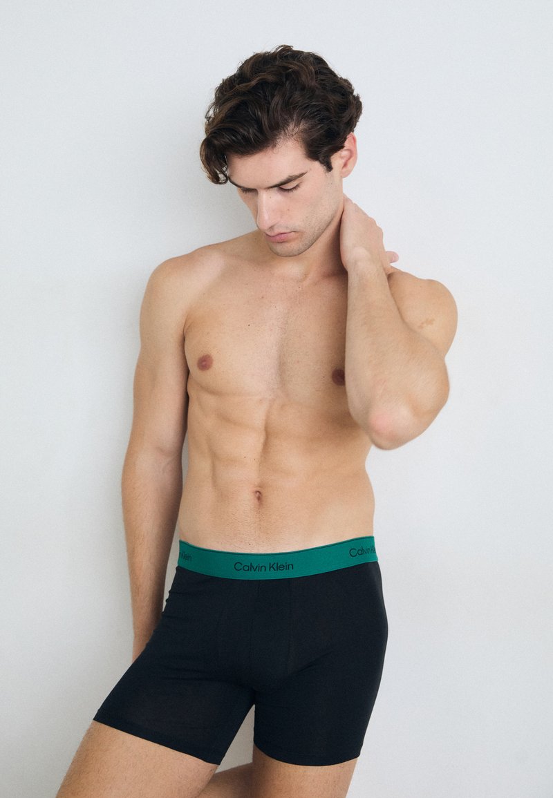 Boxer briefs neri con una cintura verde di Calvin Klein, caratterizzati da una vestibilità aderente e una texture liscia. Il modello posa su uno sfondo neutro.