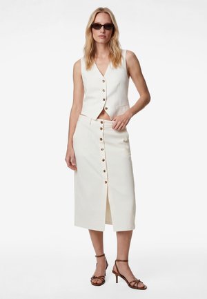 Femme portant un gilet blanc sans manches à boutons et une jupe taille haute assortie avec une fente frontale, des sandales à talons marron et des lunettes de soleil foncées.