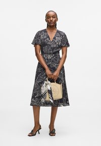 Robe noire avec des motifs de feuilles blanches, manches courtes et évasées, taille cintrée et jupe fluide. Le mannequin tient un sac à main beige avec une écharpe.