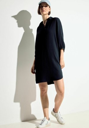 Abito nero strutturato con collo a camicia e maniche a tre quarti, abbinato a sneakers bianche e un cappellino azzurro chiaro.