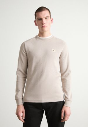 Lys beige langærmet termisk skjorte med et struktureret mønster, rund halsudskæring og et lille logo-lap på venstre bryst. Båret med mørke bukser.