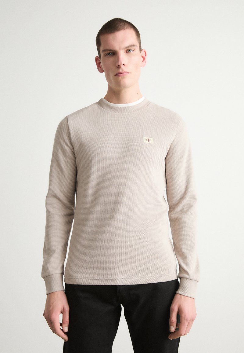 Lys beige langermet termisk skjorte med et teksturert mønster, rund halslinning og et lite logo-merke på venstre bryst. Båret med mørke bukser.