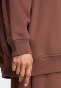 Brązowy bawełniany sweatshirt z prążkowanymi mankietami i dołem. Zbliżenie ręki opierającej się na tkaninie, podkreślające detale tekstury i dopasowania.