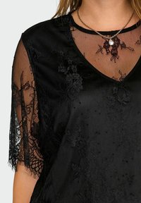 Schwarzes Spitzentop mit Blumenmustern, durchsichtige Spitzenärmel, V-Ausschnitt und goldene Kettenhalskette mit einem einzelnen weißen Perlenanhänger.