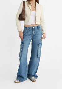 Femme portant un blazer blanc court, un crop top blanc, un jean cargo bleu à jambes larges, des baskets argentées et un sac à bandoulière marron sur fond blanc.