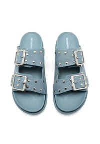 Sandalias azules hechas de material sintético que presentan dos hebillas de plata sobredimensionadas y acentos de tachuelas en la parte superior. Suela plana, diseño de punta abierta.