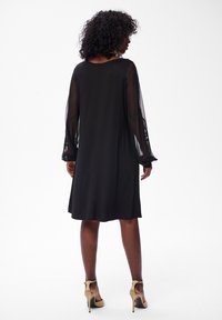Frau mit lockigem Haar, die ein knielanges schwarzes Kleid mit transparenten langen Ärmeln und beigen hohen Schuhen trägt, und vor einem weißen Hintergrund steht.