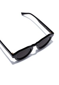 Schwarze Sonnenbrille mit runden Gläsern und einem dicken Rahmen. Das Design hat eine wellenförmige obere Kante und goldene Akzente an den Bügeln.