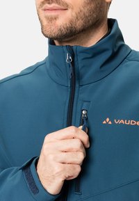 Veste bleue Vaude pour homme avec col haut et poches zippées. Présente un tissu lisse et texturé ainsi qu'un accent logo orange sur la poitrine.