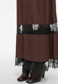 Bruine rok met doorschijnende zwarte kantafwerking. De zoom heeft een golvend ontwerp. Bruine laarzen met hak zijn gedeeltelijk zichtbaar aan de onderkant.