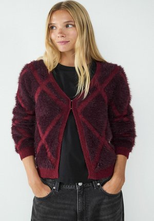 Kastanienbrauner, flauschiger Cardigan mit Rautenmuster, Reißverschluss, gerippten Bündchen und Saum. Über einem schwarzen Oberteil getragen, kombiniert mit dunklen Jeans.