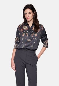 Silk blouse in dark gray with colorful floral and paisley patterns, featuring a collar and rolled-up sleeves. Paired with dark trousers.