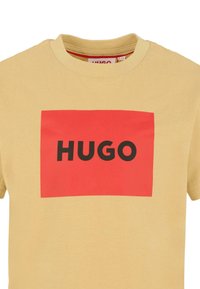 T-shirt jaune à manches courtes avec un logo rectangulaire rouge affichant le mot "HUGO" en lettres majuscules noires.