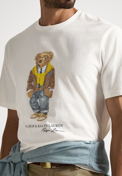 Λευκό βαμβακερό T-shirt με εικονογραφημένη αρκούδα που φορά καφέ καρό σακάκι, κίτρινο πλεκτό κασκόλ και τζιν παντελόνι. Το κείμενο από κάτω γράφει "Polo Bear by Ralph Lauren."