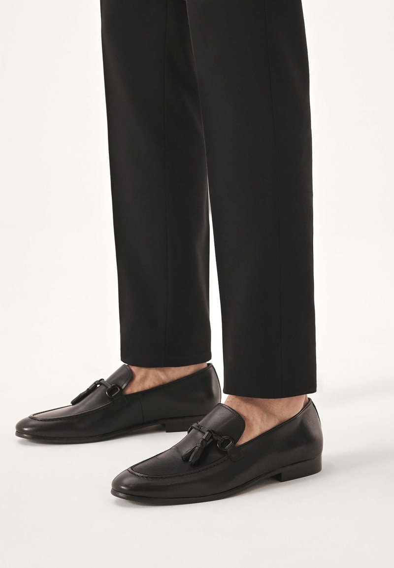 Schwarze Leder-Loafer mit glatter Oberfläche, verziert mit einer dekorativen Quaste und Metallbeschlägen, kombiniert mit maßgeschneiderten schwarzen Hosen.