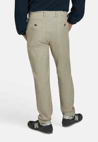 Beige broek met een gestructureerd patroon, voorzien van achterzakken met knoopsluitingen. Gekleed met zwarte sneakers en een marineblauwe trui.