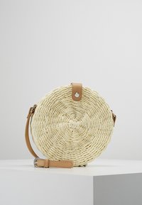 Borsa rotonda in paglia intrecciata color crema, con una tracolla in pelle marrone e design circolare con superficie testurizzata. Dettaglio in metallo nella parte superiore.