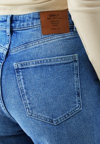 Približan posnetek zadnjega žepa modrih kavbojk iz jeansa z rjavim usnjenim označevalcem z napisom "ONLY Blue Denim, Denim Wear You Won't Forget, Anno 1995."