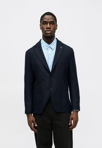 Blazer de algodón azul marino con dos bolsillos frontales, cierre de un solo botón y solapa con muesca, usado sobre una camisa de botones a rayas azul claro.