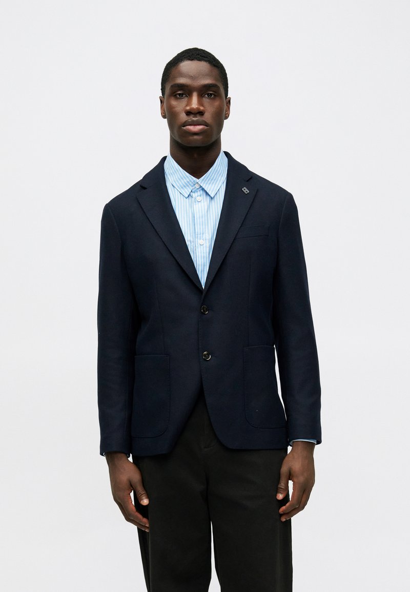 Blazer de algodón azul marino con dos bolsillos frontales, cierre de un solo botón y solapa con muesca, usado sobre una camisa de botones a rayas azul claro.