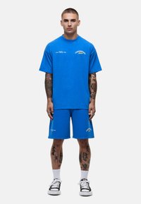 T-shirt bleu vif et shorts assortis, fabriqués en coton. Le t-shirt présente un texte blanc sur le devant ; les shorts ont un design simple avec des accents.